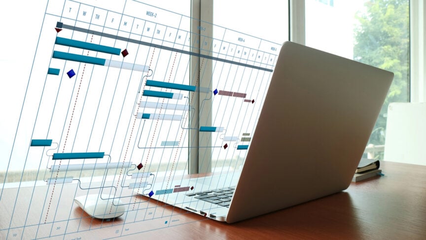 Laptop and coffee mug on desk with Gantt chart project timeline displayed on wall showing task dependencies and milestones.